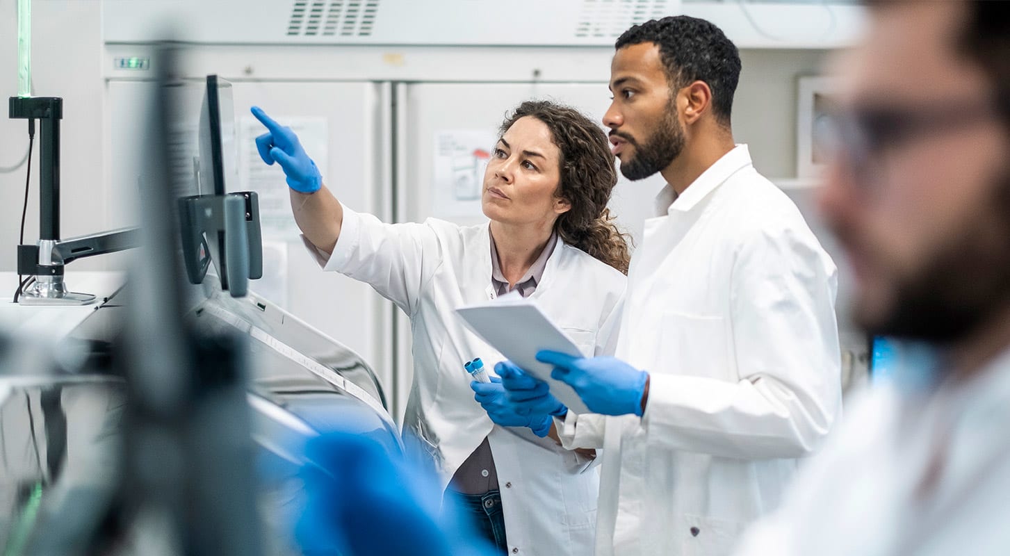 Two scientists in lab coats focused on a computer screen, discussing findings in a lab environment.